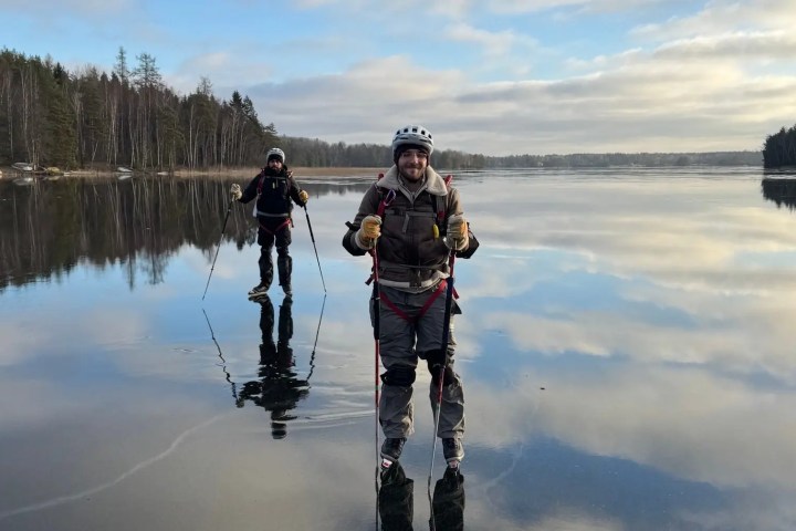 Two people ice skating on a calm frozen lake with trees in the background.